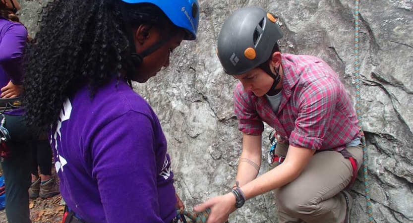 One person helps another tie a rope. Both are wearing safety gear. 